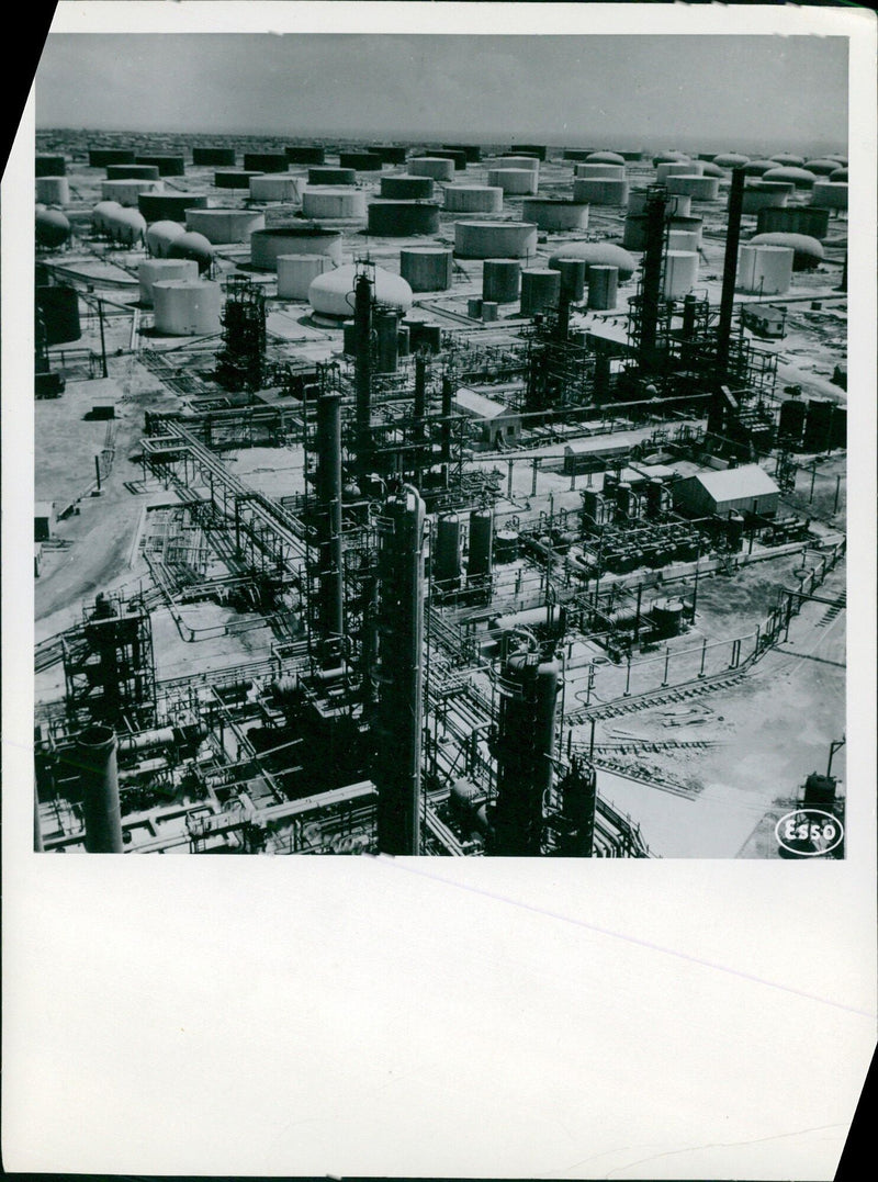 Framed by a vast network of pipes, the Esso refinery in the United States is seen with its distillation columns in the foreground and storage tanks in the background. - Vintage Photograph
