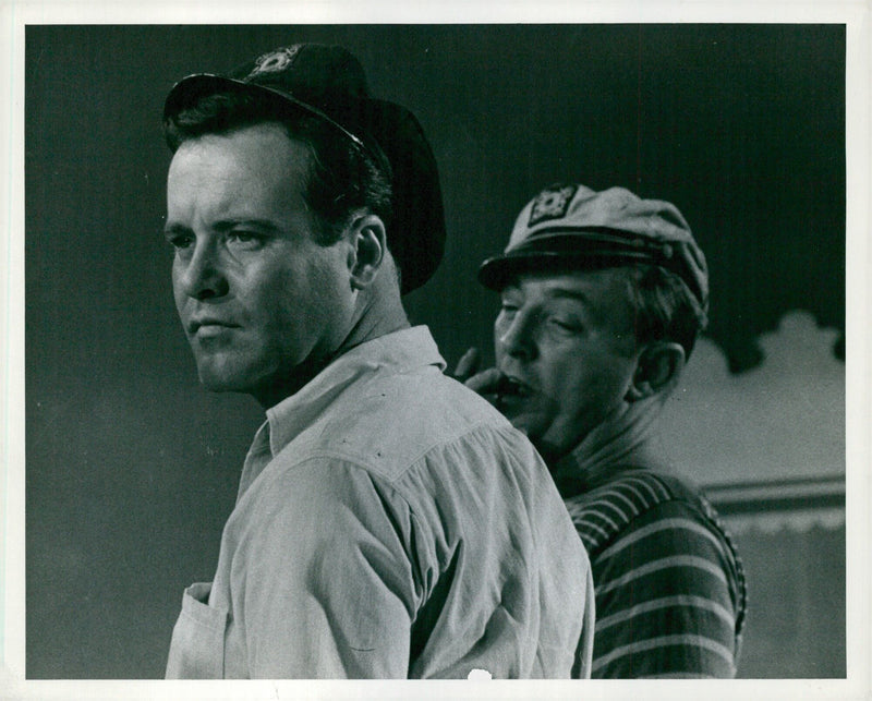 Robert Mitchum contemplates his next move while co-star Jack Lemmon looks on in a scene from the Warwick Films production of "Fire Down Below" being filmed at Elstree Studios. - Vintage Photograph