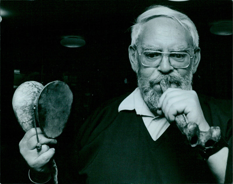 Curator Jeremy Montagu blows into a trumpet-shaped instrument made from the left thigh-bone of an old Tibetan man. - Vintage Photograph