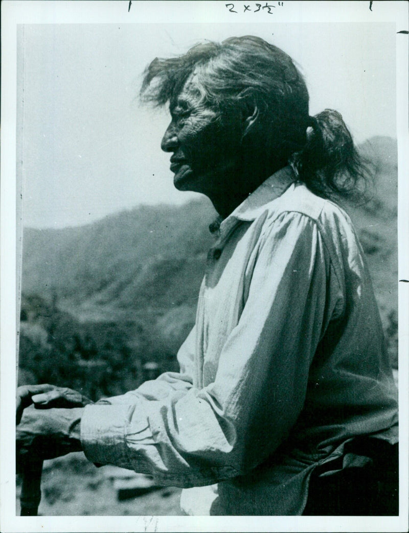 An elderly Tibetan man in traditional dress stands in front of a prayer wheel. - Vintage Photograph