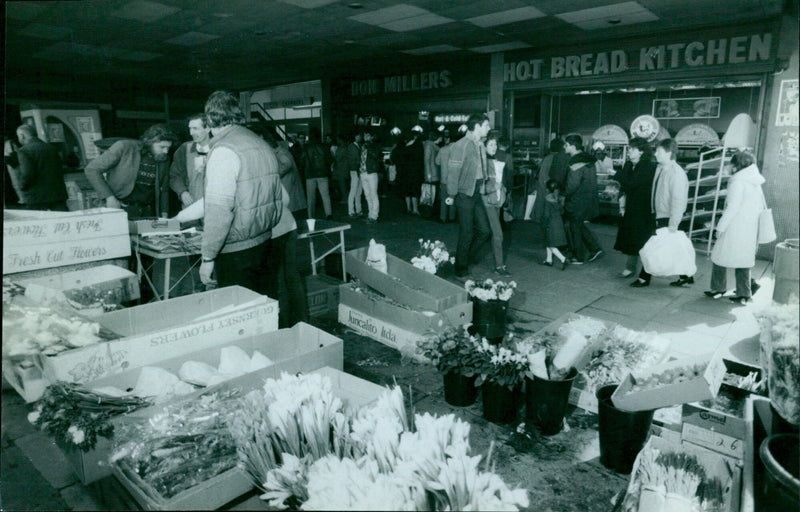 People walking in front of a flower shop, a hot bread kitchen, and the Queen Street City Chambers in Wan Ma. - Vintage Photograph