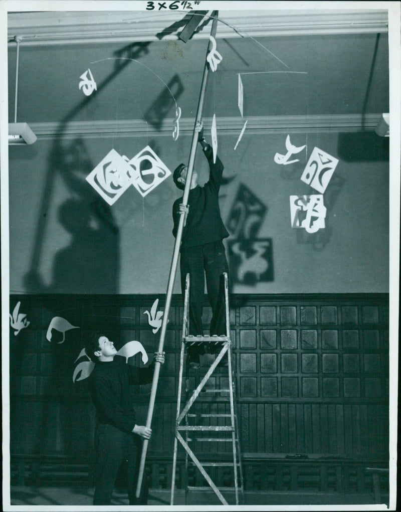 Mansfield College students John Whilley (24) and Richard Thomas (21) hang a mobile made of Eskimo artist signatures for the college ball. - Vintage Photograph
