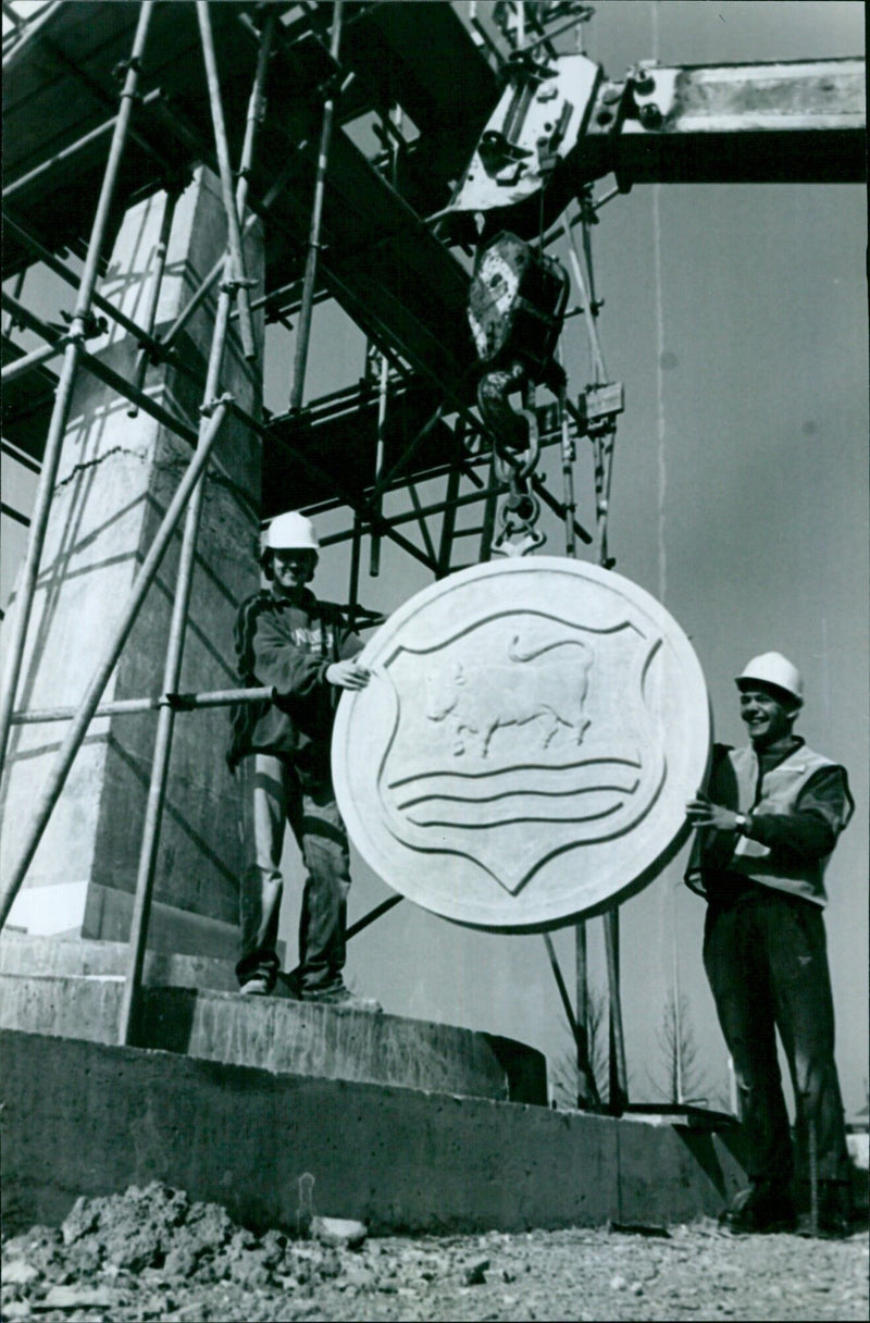 Stonemasons Danny Walker and Mick Hale lift the stone coat of arms into place at the Nuffield Needle in Oxford, England on March 23, 1995. - Vintage Photograph