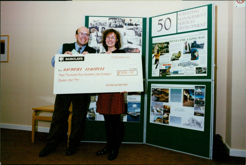 A man holds a sign showing the amount of money received by MacIntyre Adventure for a project completed by Ridge and Partners. - Vintage Photograph