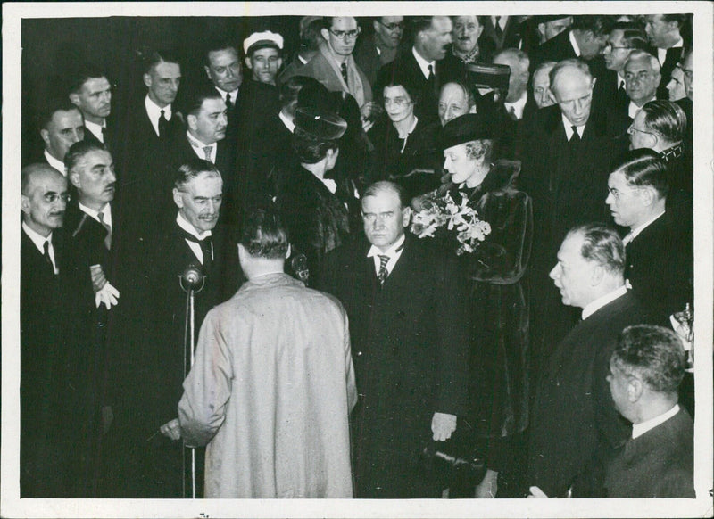 Neville Chamberlain making a statement at the microphone with M. Daladier to his left; behind them are Mrs. Chamberlain and Falifex as well as Messrs. Bonnet and Felifex. - Vintage Photograph