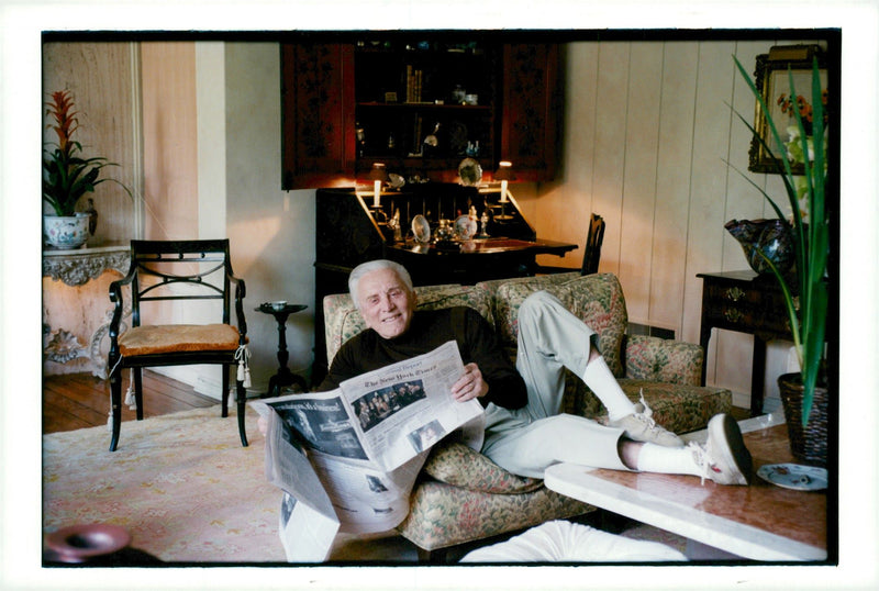 Kirk Douglas reads the newspaper on the couch in his home in Beverly Hills - Vintage Photograph
